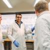 Student showing equipment in a lab wearing a lab coat and blue gloves smiling